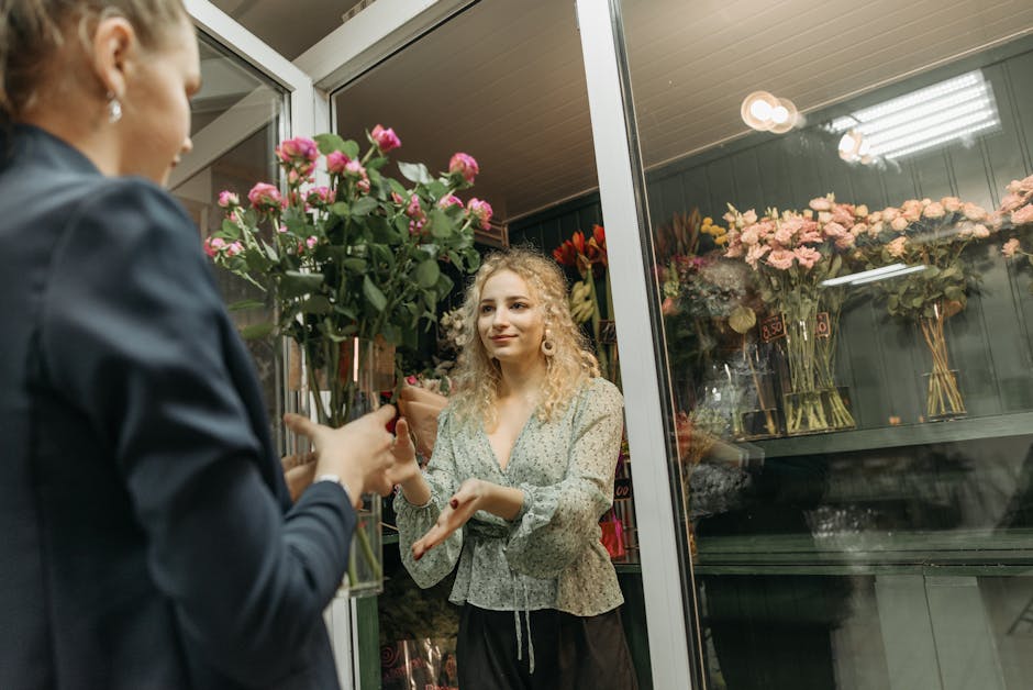 A female florist with curly blonde hair, dressed in a patterned blouse, stands inside a flower shop next to a glass display case filled with bouquets of pink roses and other mixed flowers. The florist