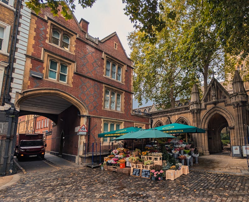 A vibrant flower stall set up under a large green umbrella in Stoke Newington Church Street, displaying an assortment of fresh flowers including pink peonies, white roses, yellow tulips, and mixed gre