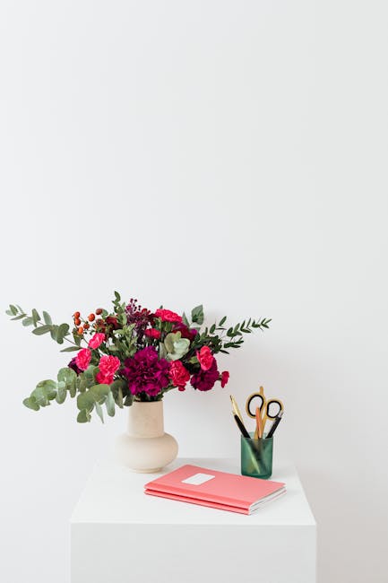 A fresh floral arrangement featuring deep pink and crimson carnations, fuchsia gerbera daisies, and silver-green eucalyptus leaves arranged in a matte beige ceramic vase, placed on a white surface aga