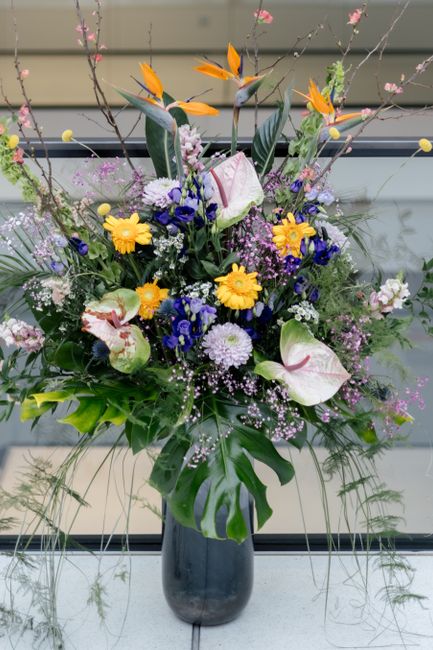 A vibrant, mixed floral arrangement in a tall, dark glass vase featuring large white anthuriums with pink-tipped edges, yellow daisies, deep blue delphiniums, and purple asters. The bouquet includes s